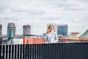Una giovane donna d'affari con smartphone e laptop in piedi su una terrazza, facendo una telefonata quando lavora.