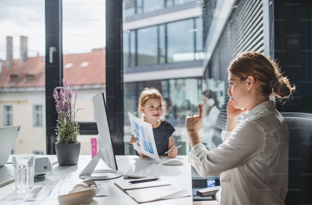 Uma empresária com uma filha pequena sentada em um escritório, trabalhando.