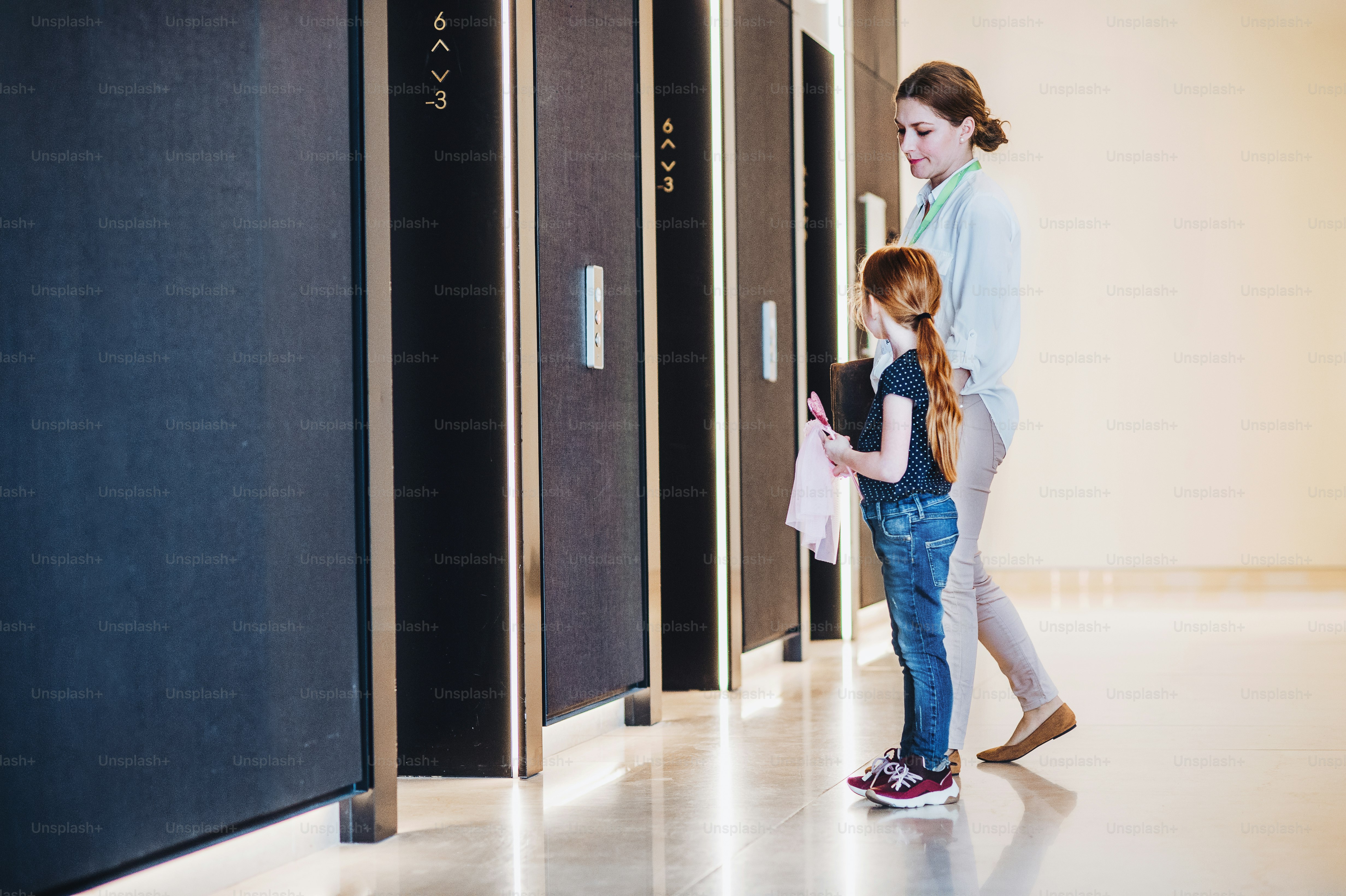 A side view of businesswoman with small daughter standing by elevators in office building.