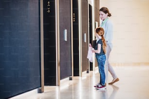 Uma vista lateral de empresária com filha pequena de pé por elevadores no prédio de escritórios.