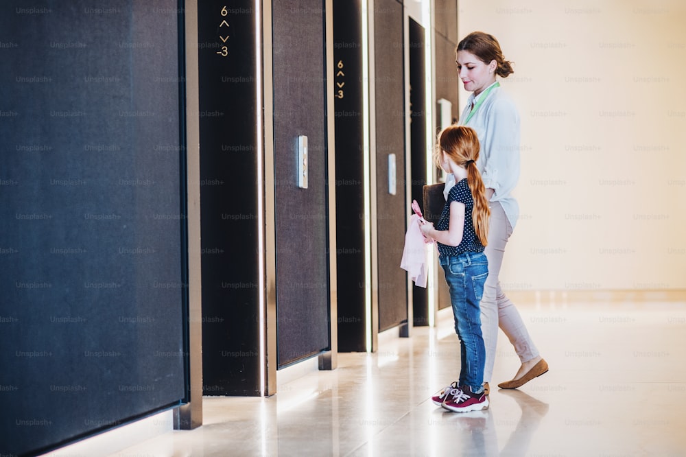 Eine Seitenansicht einer Geschäftsfrau mit kleiner Tochter, die an Aufzügen im Bürogebäude steht.