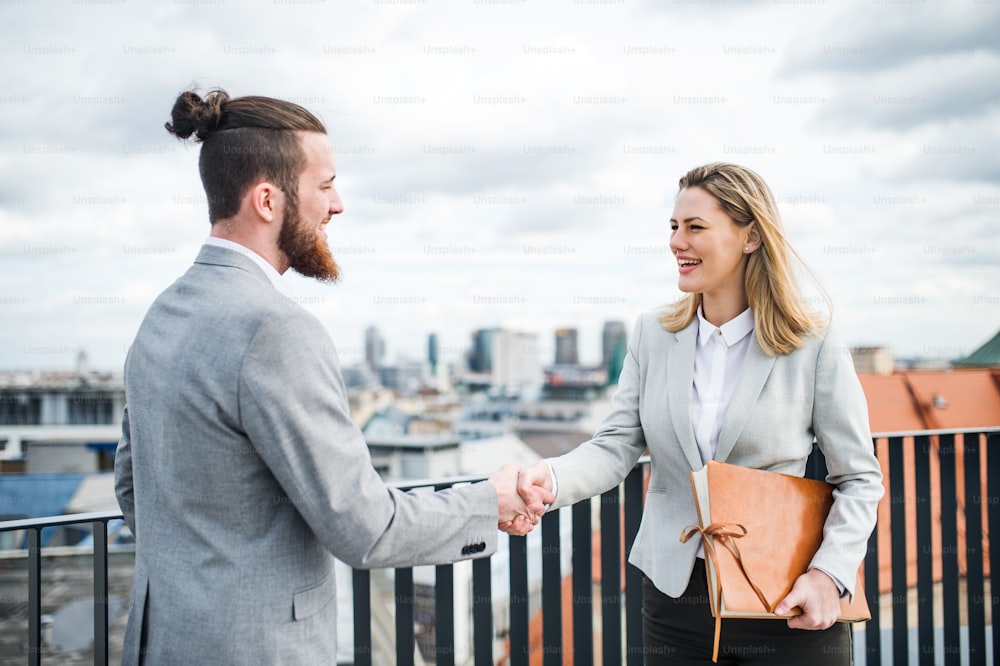 Two young businesspeople standing on a terrace outside office, shaking hands.