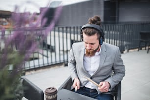 A young businessman with headphones and drumsticks sitting on a terrace, having fun.