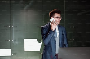 A portrait of young businessman with smartphone standing by lockers in an office, making a phone call. Copy space.
