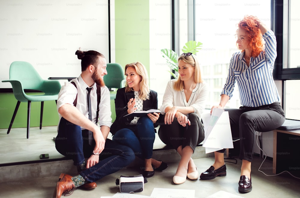 A group of young business people with a diary sitting on the floor in an office, talking.