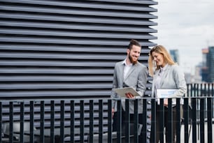 Dos jóvenes empresarios alegres con tableta de pie en una terraza fuera de la oficina, trabajando.