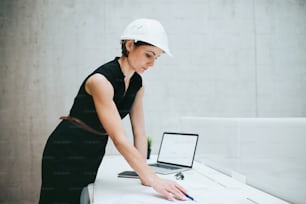 Una joven empresaria o arquitecta con casco y computadora portátil de pie en el escritorio de la oficina.