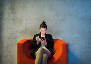 Une jeune femme d’affaires avec smartphone assise sur un fauteuil rouge dans le bureau, un mur de béton gris en arrière-plan.