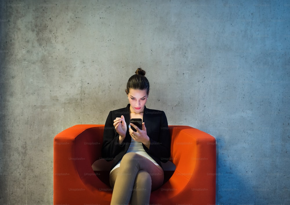A young businesswoman with smartphone sitting on red armchair in office, a gray concrete wall in the background.