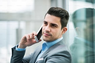 Un ritratto di giovane uomo d'affari felice con lo smartphone seduto nel corridoio fuori dall'ufficio, facendo una telefonata.