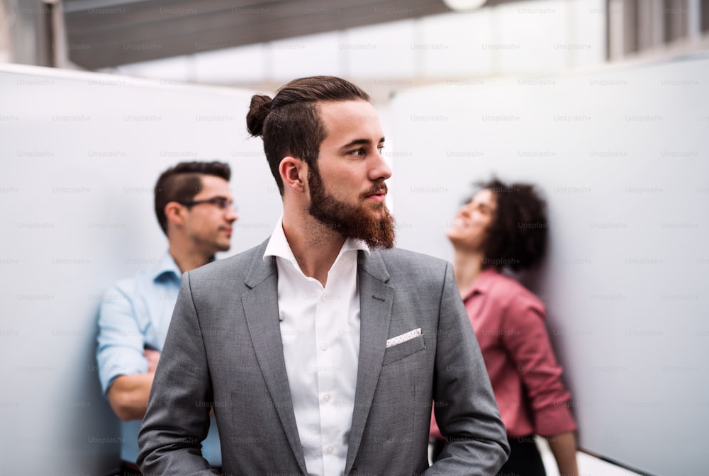 Un retrato de un joven hombre de negocios de traje de pie en la oficina, con colegas en el fondo.