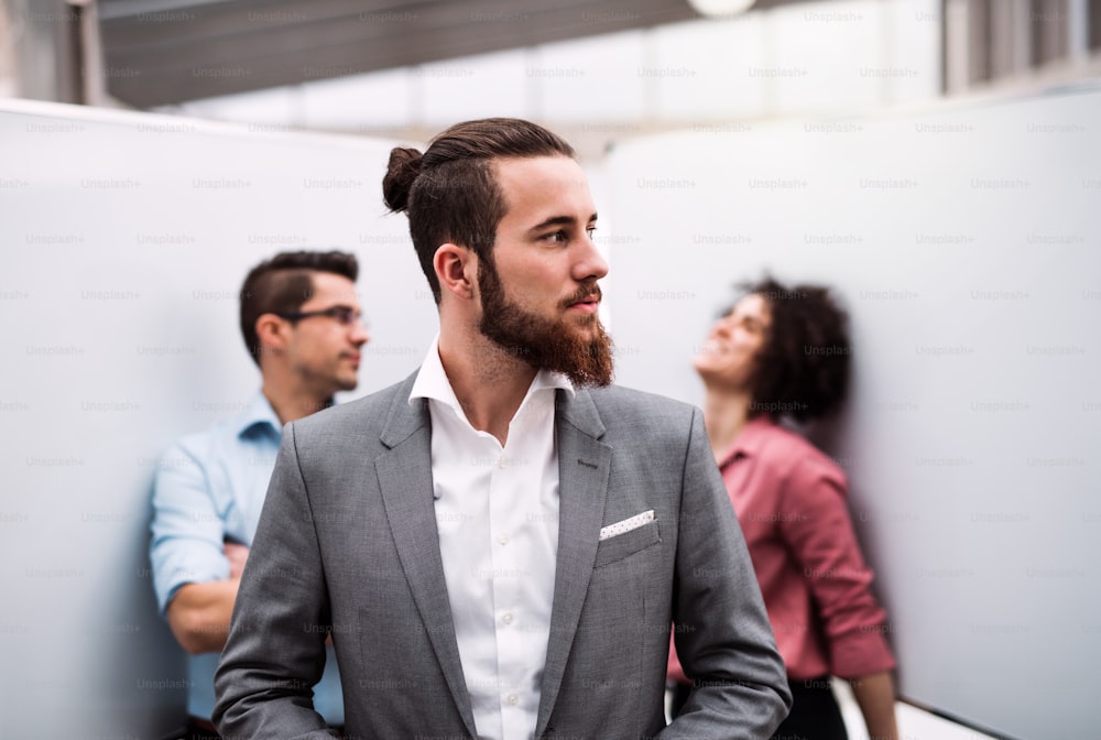 A portrait of young businessman in suit standing in office, colleagues in the background.