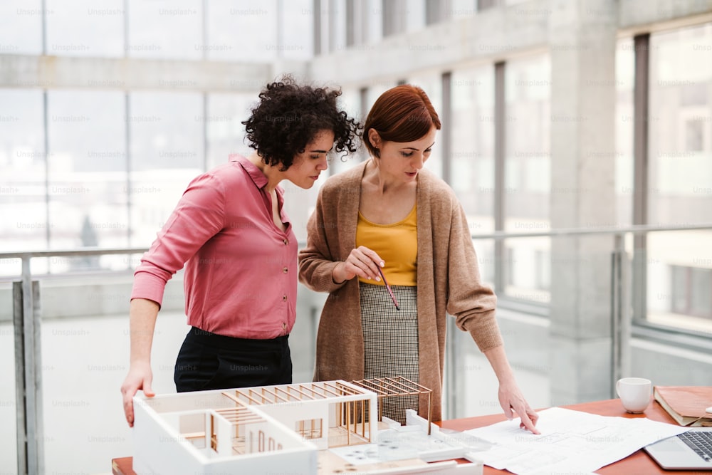 Dos jóvenes arquitectas con el modelo de una casa de pie en la oficina, trabajando y hablando.