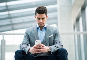 Ein Porträt eines glücklichen jungen Geschäftsmannes mit Smartphone, der auf dem Flur vor dem Büro sitzt.