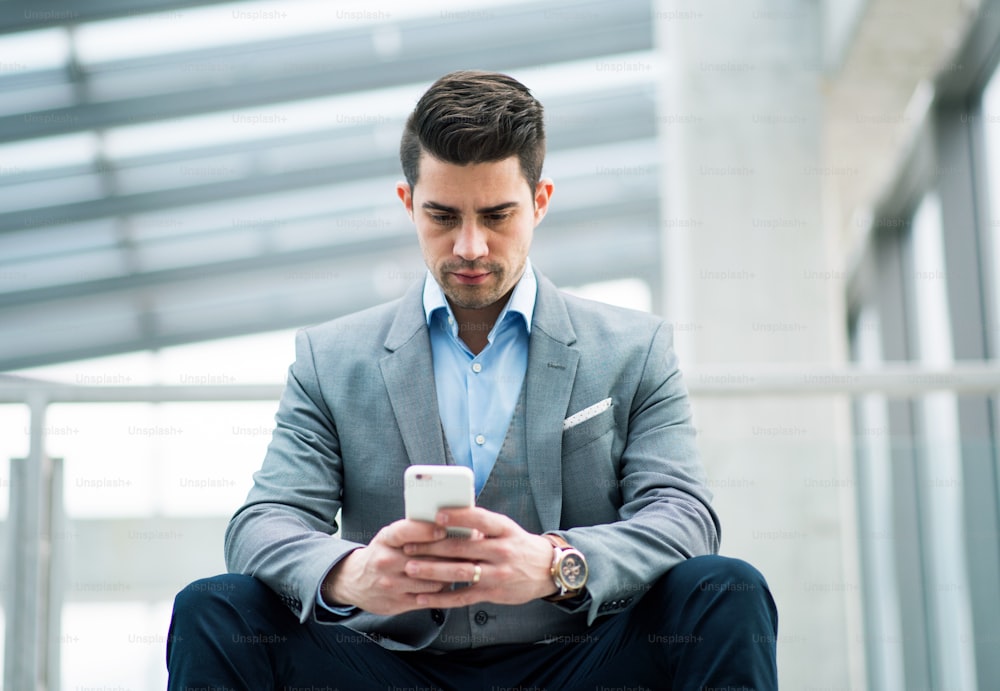 オフィスの外の廊下に座っているスマートフォンを持つ幸せな若いビジネスマンのポートレート。