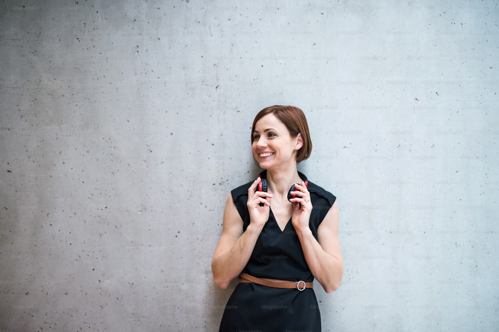 A portrait of young business woman with headphones standing against concrete wall in office.