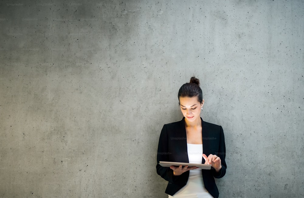 Um retrato de uma jovem empresária feliz com tablet digital de pé contra a parede de concreto no escritório.