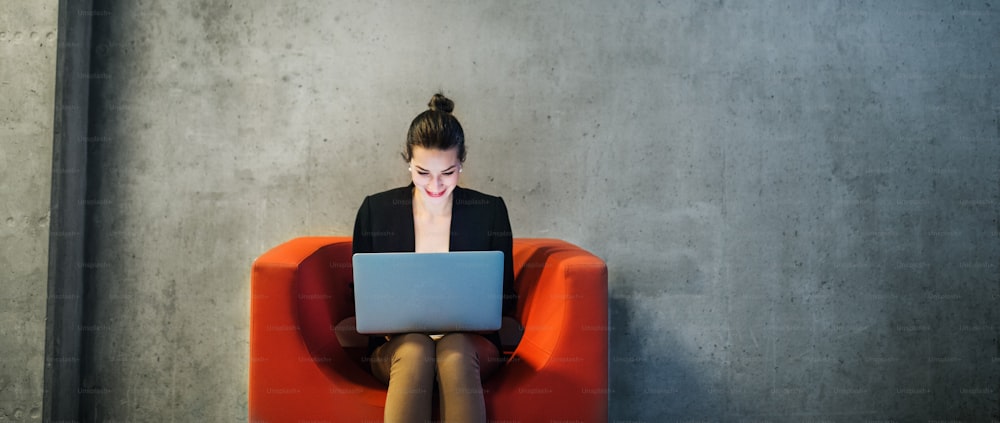 Una giovane donna d'affari con computer portatile seduta su poltrona rossa in ufficio, un muro di cemento grigio sullo sfondo. Copia spazio.