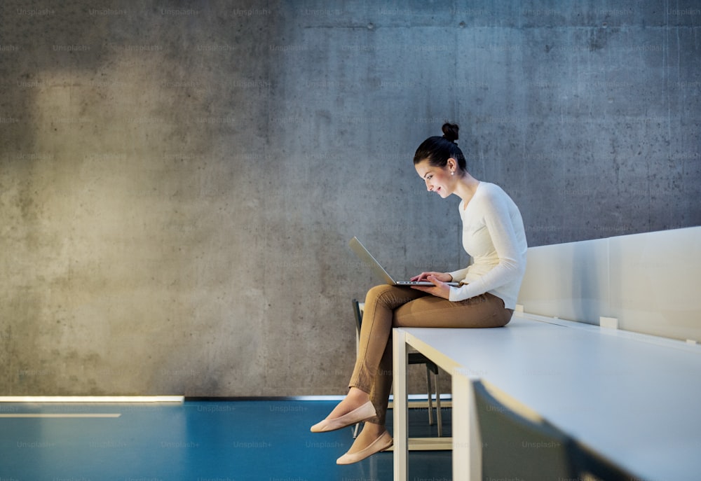 Portrait d’une jeune étudiante ou femme d’affaires assise sur un bureau dans une pièce d’une bibliothèque ou d’un bureau, à l’aide d’un ordinateur portable.
