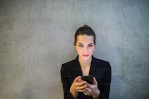A front view of happy young business woman with smartphone standing against concrete wall in office.