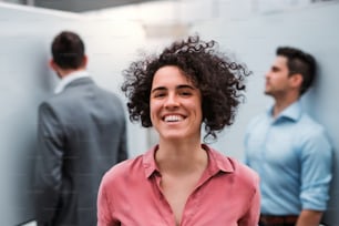 A portrait of young businesswoman standing in office, colleagues in the background.