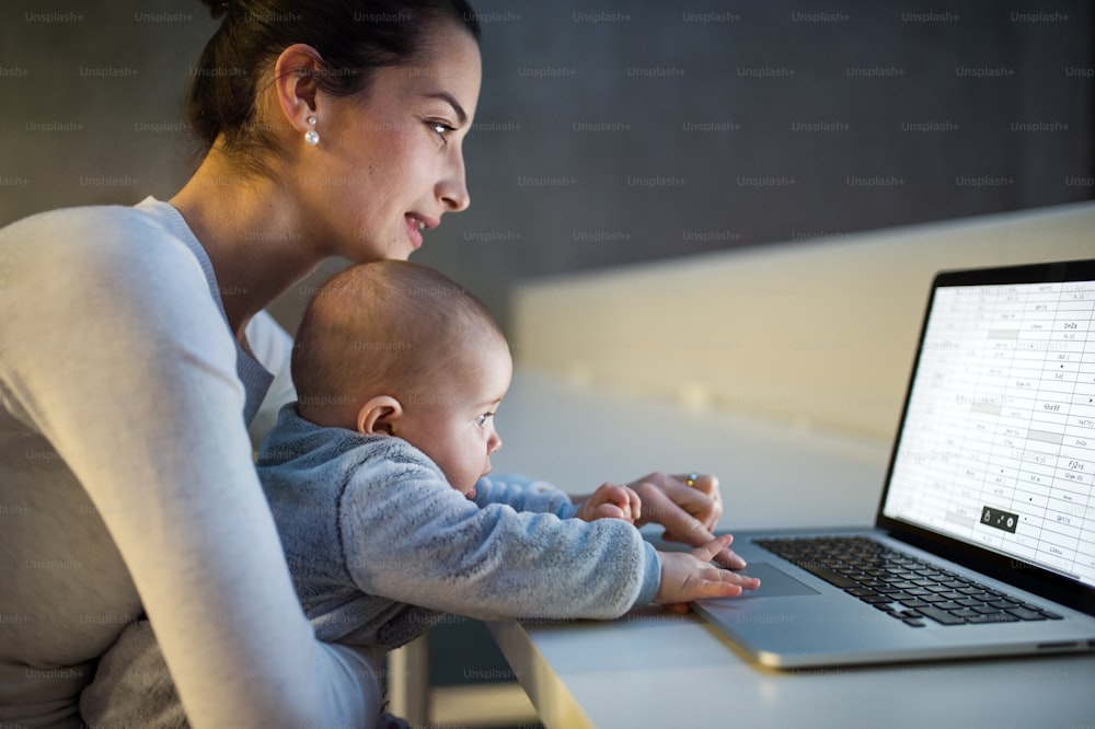 A side view of a young student mother or businesswoman sitting on desk with a baby in room in a library or office, using laptop.