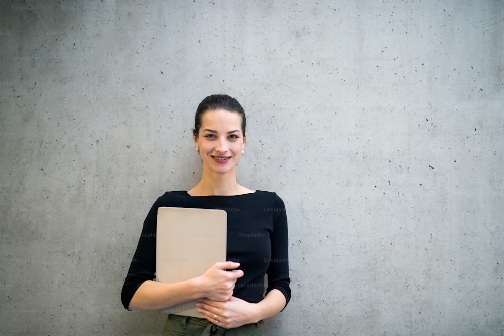 A portrait of happy young business woman with clipboard standing against concrete wall in office. Copy space.