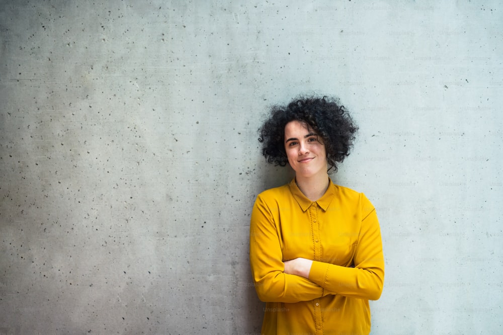 A portrait of a young student or businesswoman standing in room in a library or office, arms crossed.