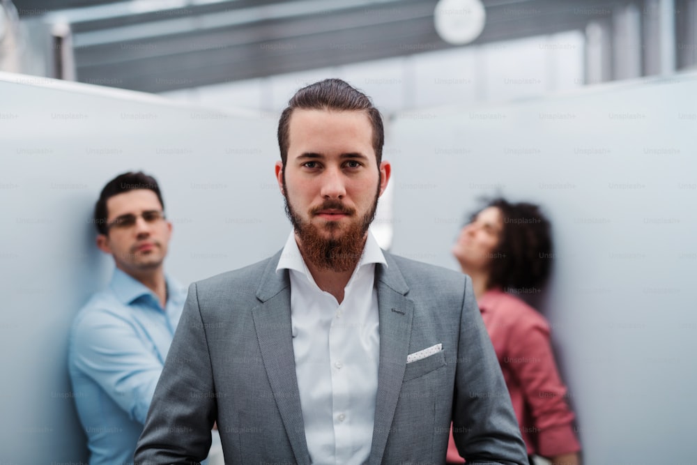 A portrait of young businessman in suit with colleagues standing in office.