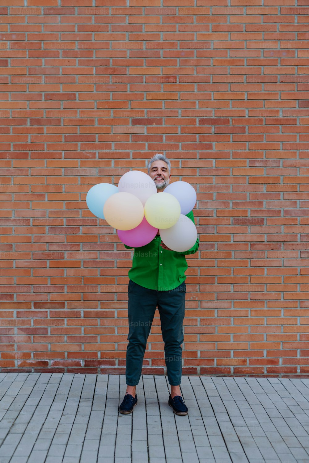 A fun portrait of happy energetic mature man holding balloons in street and hiding behind them, feeling free.