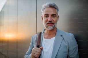 A mature relaxed businessman leaning on street wall and looking at camera, work life balance concept.