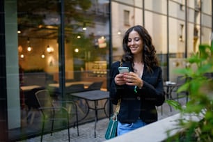 Una donna felice che invia messaggi al cellulare e aspetta fuori dal caffè in città.