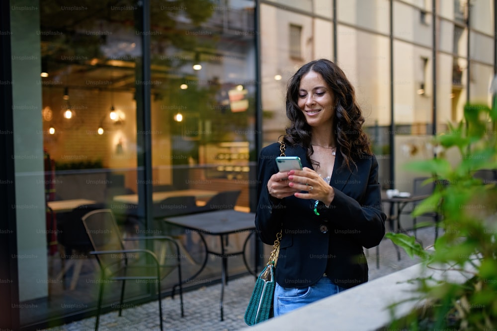 휴대 전화로 문자 메시지를 보내고 도시의 카페 밖에서 기다리는 행복한 여성.