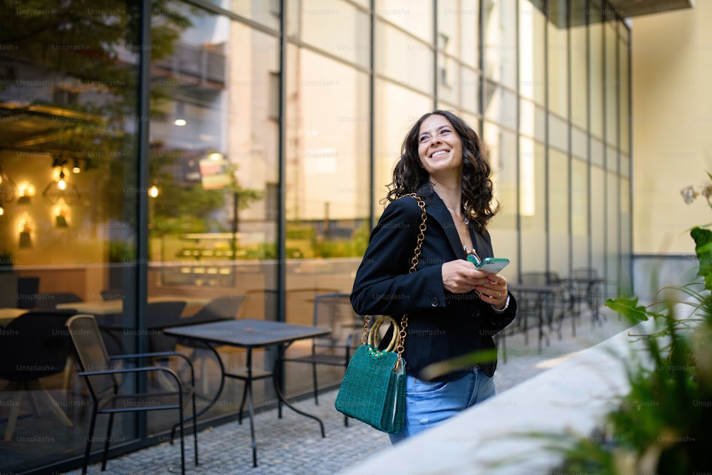 Uma mulher feliz enviando mensagens de texto no celular e esperando do lado de fora do café na cidade.