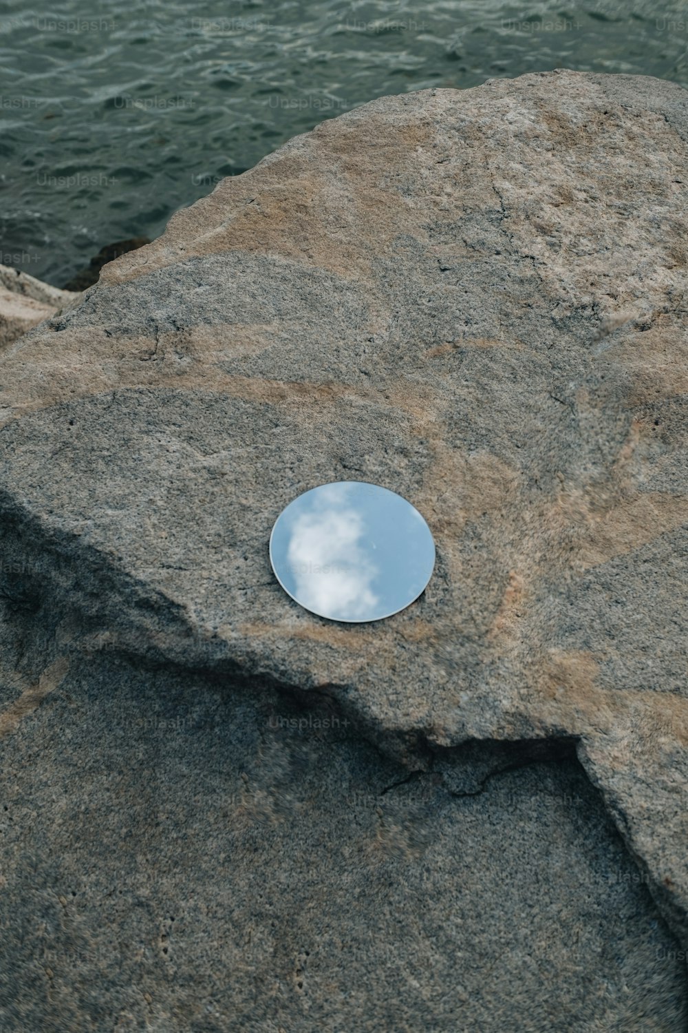 a round mirror sitting on top of a rock next to the ocean