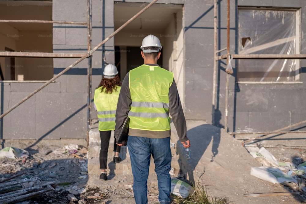 a man and a woman in safety vests walk through a construction site