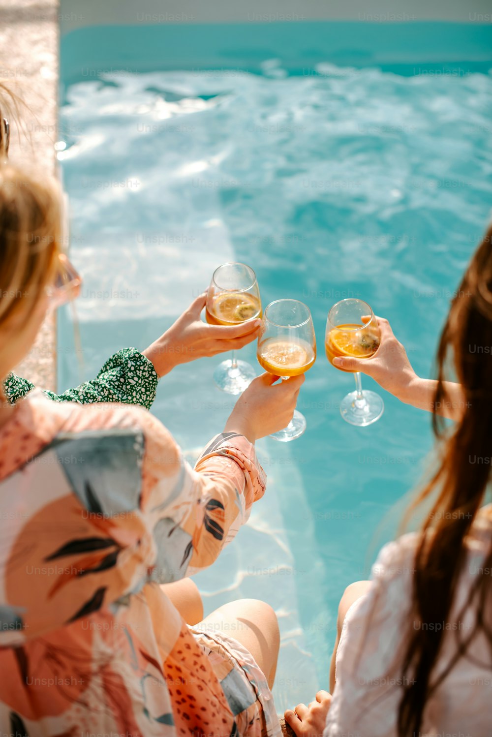 a group of women sitting next to a swimming pool holding wine glasses