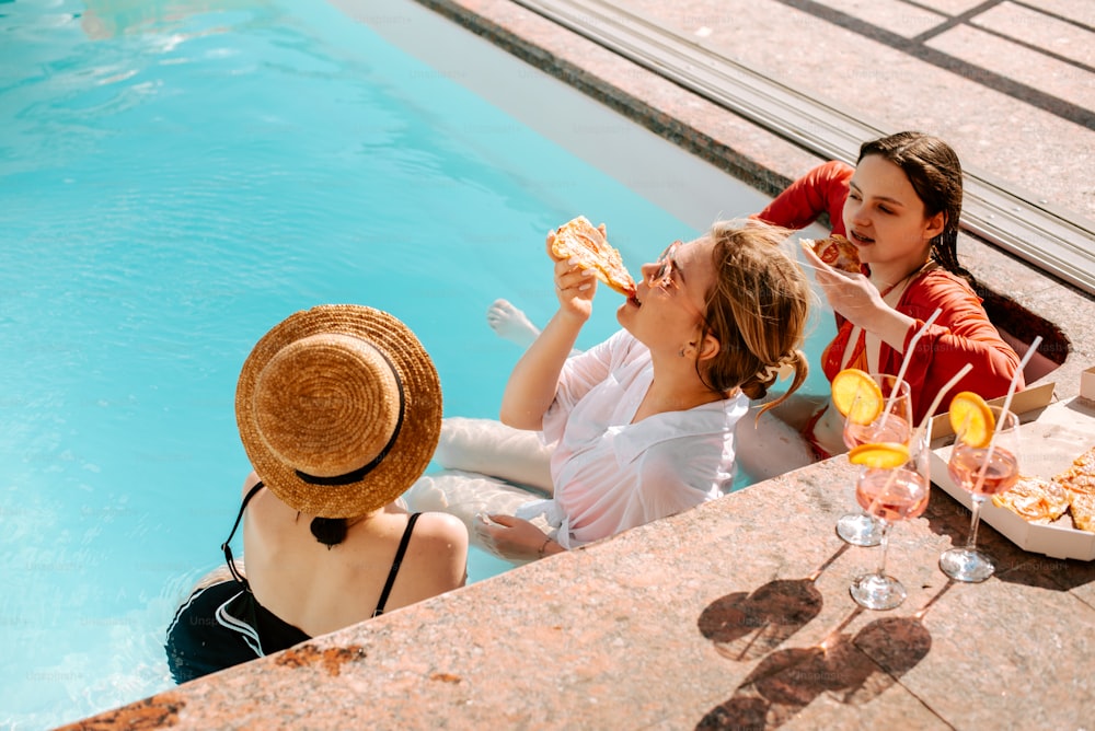 a group of women sitting next to a pool eating pizza