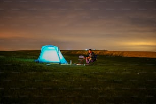 a man sitting next to a tent on top of a lush green field