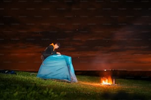 a man sitting on top of a blue tent next to a fire