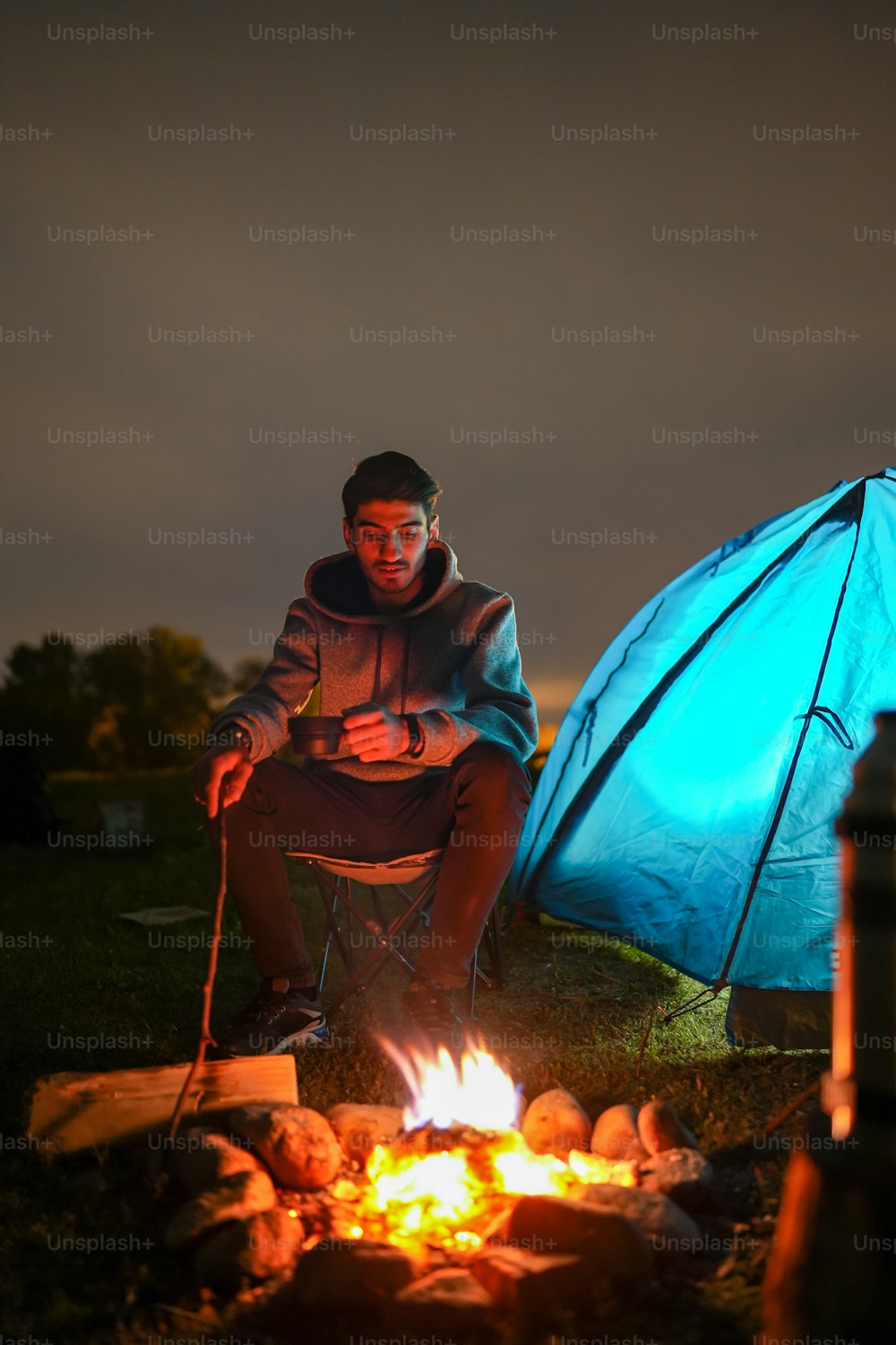파란 텐트 옆 모닥불 앞에 앉아 있는 남자