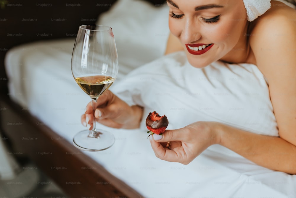 한 여자가 와인 한 잔과 과일 한 조각을 들고 침대에 누워 있다