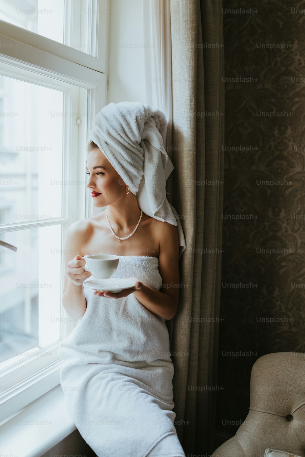 Una mujer con una toalla sosteniendo una taza y mirando por una ventana