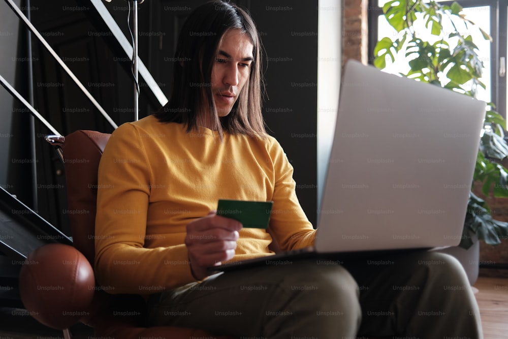 Un hombre sentado en una silla sosteniendo una tarjeta de crédito y una computadora portátil