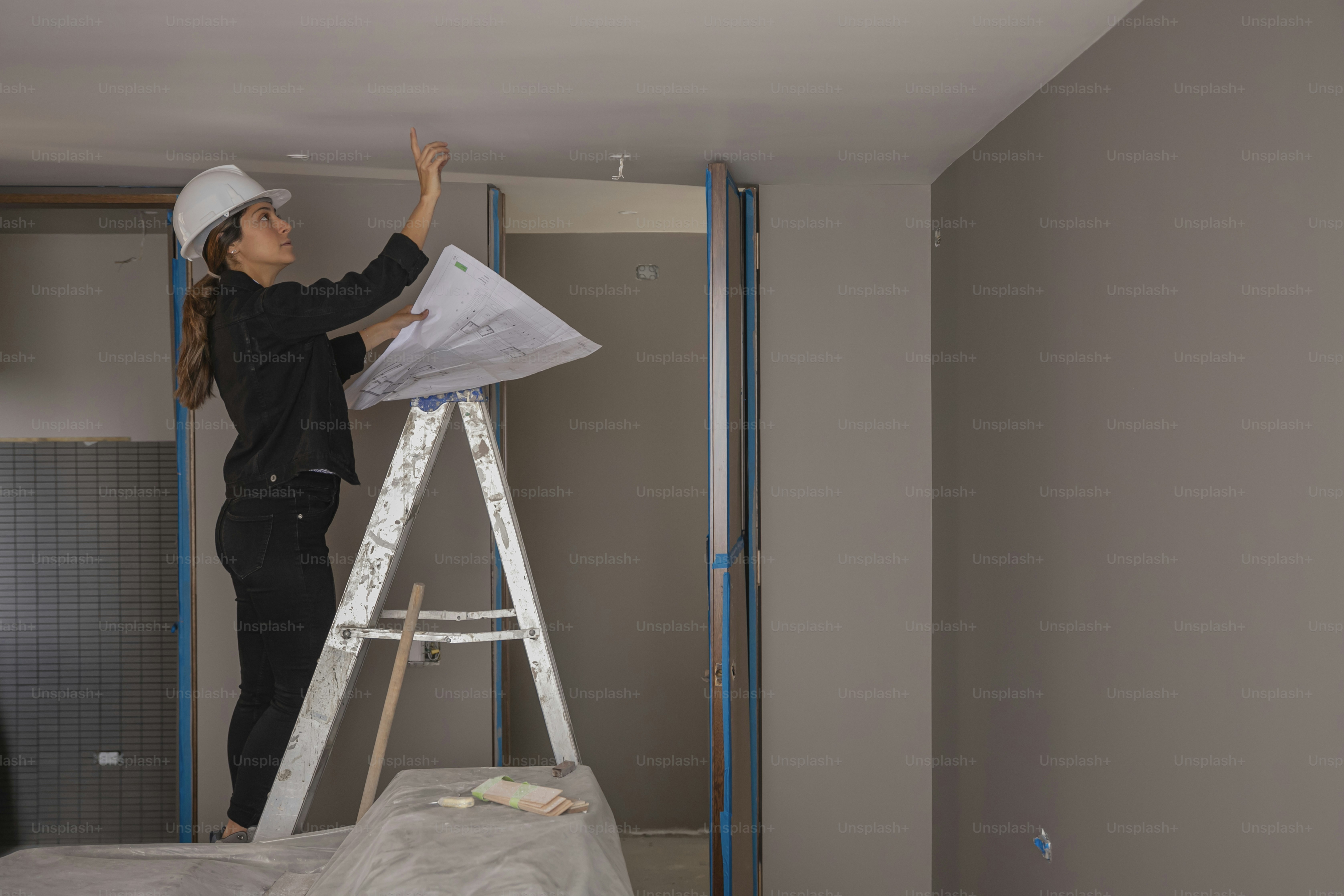 Frau mit Schutzhelm auf Leiter inspiziert die Decke