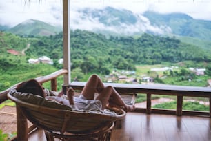 Una giovane donna che tiene la tazza di caffè mentre è sdraiata sul divano sul balcone, guardando le montagne e la natura verde