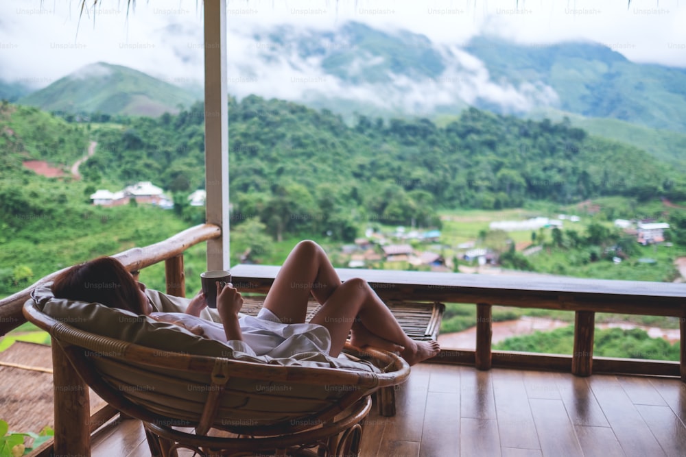 A young woman holding coffee cup while lying on sofa on balcony , looking at mountains and green nature