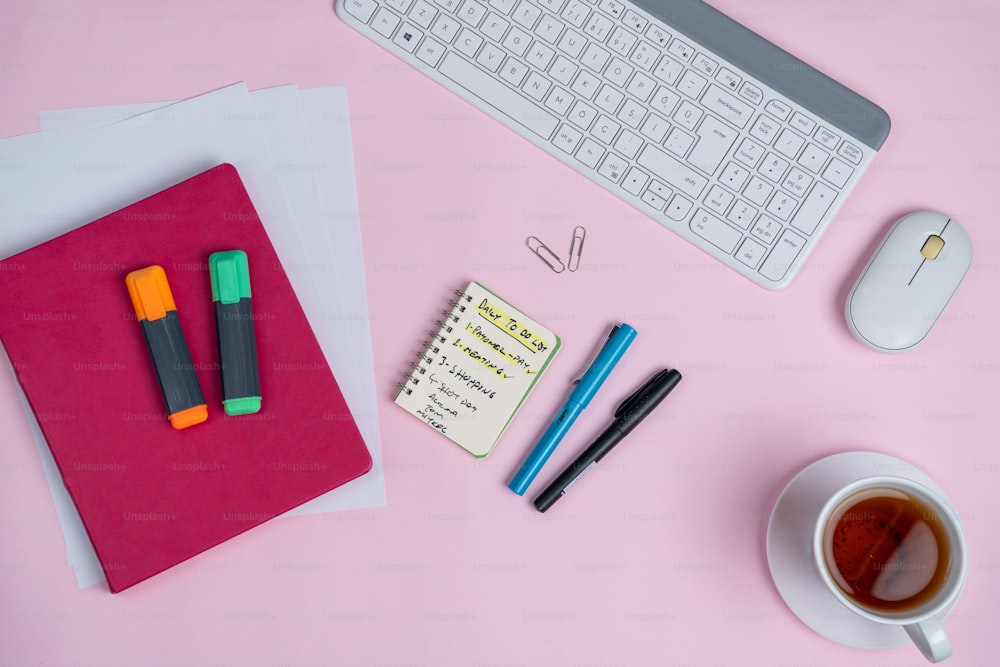 Un escritorio rosa con teclado, cuaderno, ratón y una taza de café