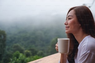 Portrait image of a beautiful asian woman holding and drinking hot coffee while looking at mountains and green nature on foggy day