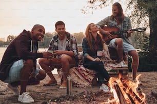 Gruppe junger Leute in Freizeitkleidung, die lächeln, während sie eine Strandparty in der Nähe des Lagerfeuers genießen